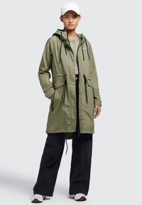 Jachetă ușoară de culoare verde olive, cu glugă ajustabilă, șnururi la talie și închidere pe bază de capse. Purttată peste un top scurt și pantaloni negri.