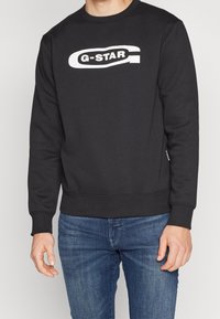 Černá mikina G-Star s bílým logem na hrudi, nošená osobou stojící v modrých džínách, oříznutá u krku a stehen.