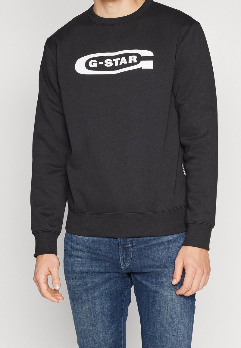 Černá mikina G-Star s bílým logem na hrudi, nošená osobou stojící v modrých džínách, oříznutá u krku a stehen.