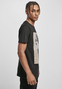 Schwarzes Baumwoll-T-Shirt mit einem frontalen Grafikdruck eines Porträts auf einem beigefarbenen strukturierten Hintergrund, mit kurzen Ärmeln und Rundhalsausschnitt.