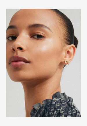 Złote kolczyki z misternymi wzorami i teksturowanym wykończeniem. Skóra modelki naturalnie się świeci, co doskonale komponuje się z biżuterią.