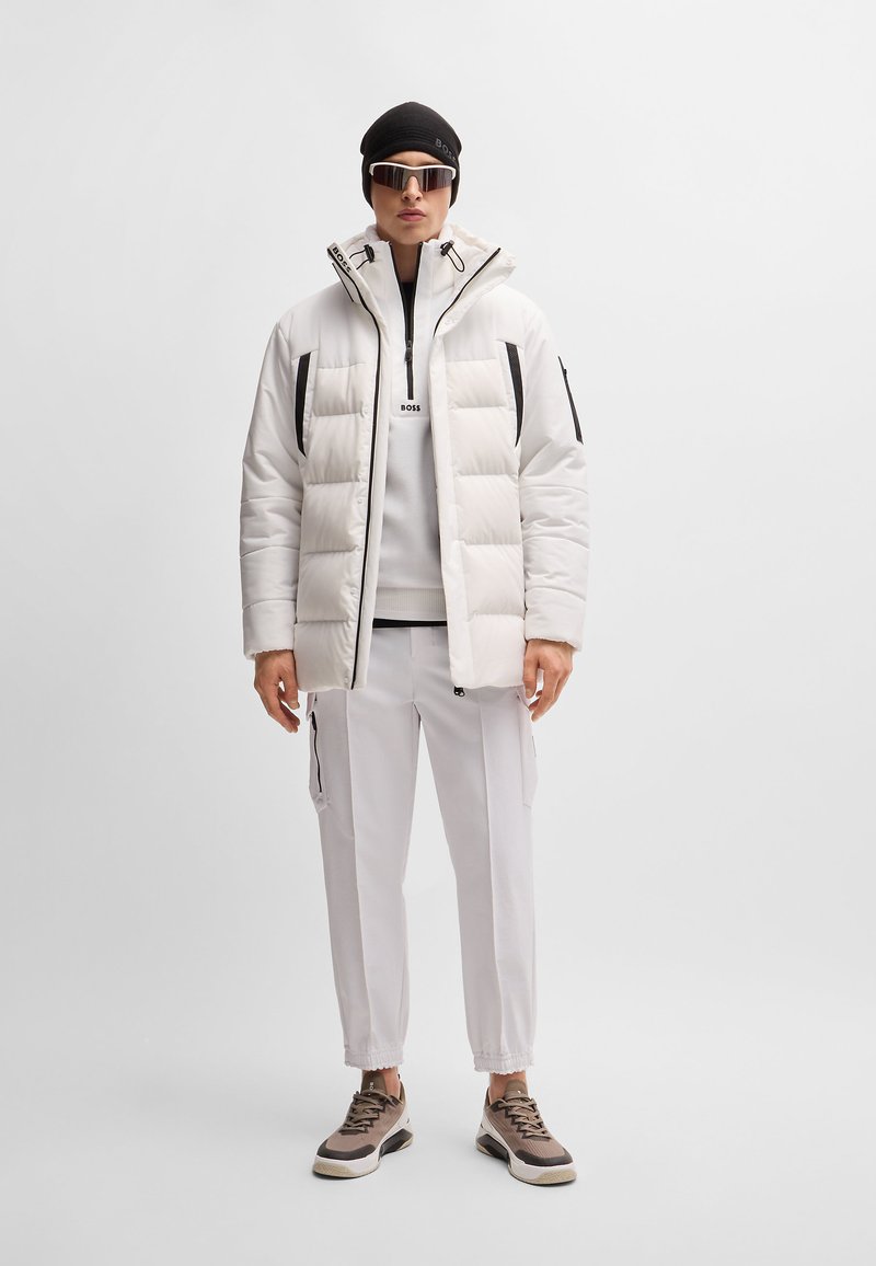 Weiße Pufferjacke mit schwarzen Akzenten, mit Reißverschlusstaschen, graue Mittelschicht, helle Hose mit Reißverschlüssen an den Seiten und sportliche braune Sneakers.