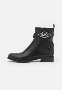 Botte de cheville en cuir noir avec un talon bas, sangle décorative et boucle circulaire en argent avec le logo "MK" sur le côté.