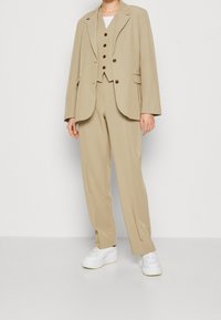 Personne portant un costume beige avec une veste, un gilet boutonné, un pantalon assorti et des baskets blanches se tenant devant un fond blanc.