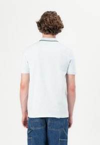 Mladý človek so svetlo hnedými kučeravými vlasmi, oblečený v svetlomodrej polokošeli a modrých rifľových cargo kraťasoch, stojaci chrbtom k jednoliatemu bielemu pozadiu.