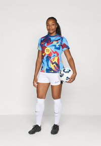 Nike uniform piłkarski w niebieskim kolorze z wielokolorowymi abstrakcyjnymi wzorami i logiem, w zestawieniu z białymi szortami i czarnymi korkami, trzymający piłkę.