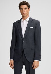 Donkergrijs op maat gemaakt colbert met een subtiel ruitpatroon, sluiting met één knoop en twee voorzakken. Wit shirt eronder, effen achtergrond.