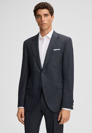 Donkergrijs op maat gemaakt colbert met een subtiel ruitpatroon, sluiting met één knoop en twee voorzakken. Wit shirt eronder, effen achtergrond.