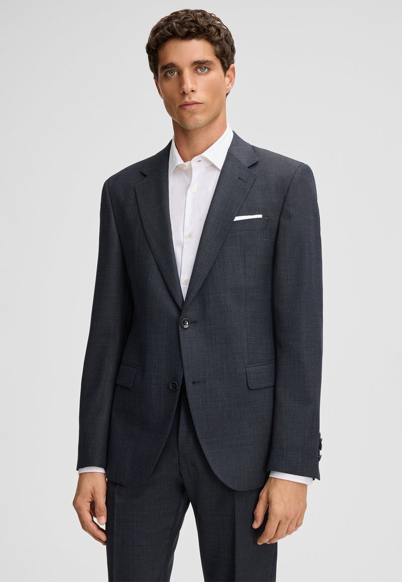 Donkergrijs op maat gemaakt colbert met een subtiel ruitpatroon, sluiting met één knoop en twee voorzakken. Wit shirt eronder, effen achtergrond.