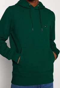 Sudadera con capucha verde hecha de una mezcla de algodón, que cuenta con un bolsillo frontal, cordones metálicos y un pequeño logo en el lado izquierdo del pecho.