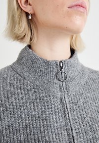 Pull en tricot gris avec col haut, doté d'une fermeture éclair et d'une tirette circulaire. La texture est côtelée et douce.