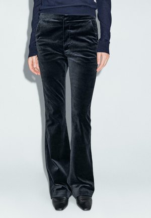 Pantalons évasés en velours noir avec une texture lisse et des poches avant, associés à un pull ajusté bleu marine et des talons noirs.