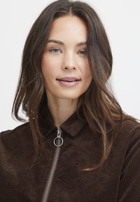 Hnedá kordrová bunda so zipsom a okrúhlym ťahadlom. Má golier a textúrovaný povrch s viditeľným ryhovaním.