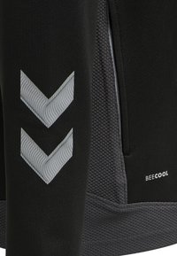 Pantalons de sport noirs avec un tissu texturé au bas, des accents en chevron argentés et une fermeture éclair sur le côté. Le logo de la marque "BECOOL" est visible.