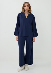 Marineblaues Zweiteiler-Outfit mit einem lockeren, langärmligen Oberteil mit V-Ausschnitt und weit geschnittenen Hosen. Hergestellt aus einem weichen, strukturierten Stoff.