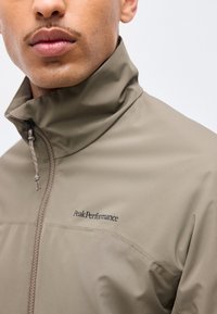 Νεαρός άνδρας που φοράει ένα ταφ τζάκετ με φερμουάρ και ψηλό γιακά, με το λογότυπο "Peak Performance" στο στήθος.