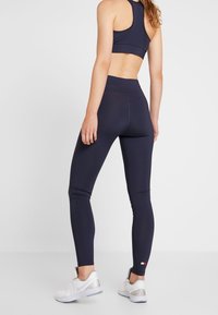 Marinblå träningsleggings med hög midja, elegant design och kontrasterande logotyp vid vristen, i kombination med vita sportskor.