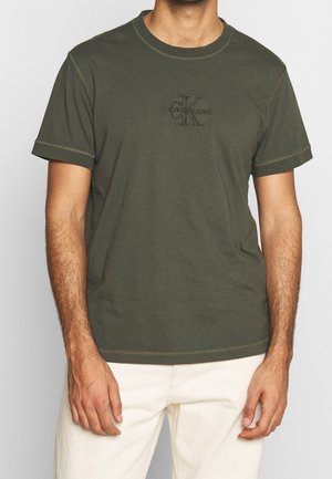 Homme portant un t-shirt Calvin Klein Jeans à manches courtes vert olive et un pantalon blanc cassé, debout avec les bras détendus le long du corps.