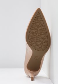 Chaussure à talon haut couleur nude avec un bout pointu, une finition en vernis lisse et une semelle marron texturée arborant un motif de logo embossé.