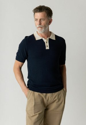 Mand med gråt skæg iført marineblå kortærmet strikket polo og beige bukser, stående med hænderne i lommerne, kiggende til venstre.