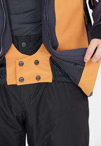 Farvet opdelt overtøj har orange og marineblå paneler med lynlås, trykknapper og elastisk talje. Materialet er struktureret og holdbart.
