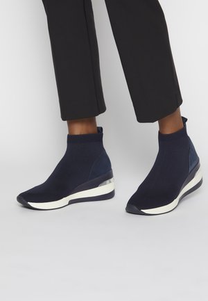 Personne portant un pantalon court noir et des baskets compensées style chaussettes bleu foncé avec des semelles blanches sur un fond uni.