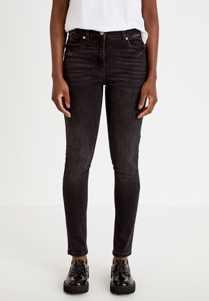 Person trägt eng anliegende schwarze Jeans, weißes Hemd, das eingesteckt ist, und glänzende schwarze Schnürschuhe, steht vor einem schlichten weißen Hintergrund.