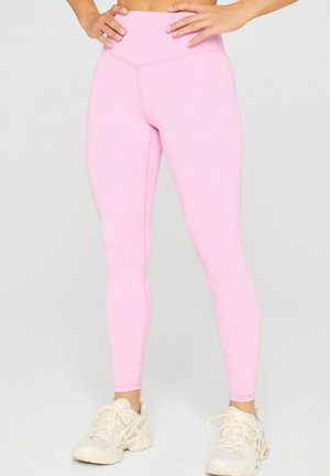 Person wearing high-waisted light pink leggings and white running shoes, standing with hands resting on hips against plain background.