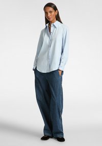 Camisa abotonada de color azul claro con cuello, mangas largas y detalles en botones plateados. Combinada con pantalones de mezclilla oscuros de pierna ancha con acentos laterales.