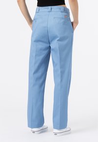 Person wearing light blue straight-leg pants and white sneakers, standing with hands in pockets, against a plain white background.