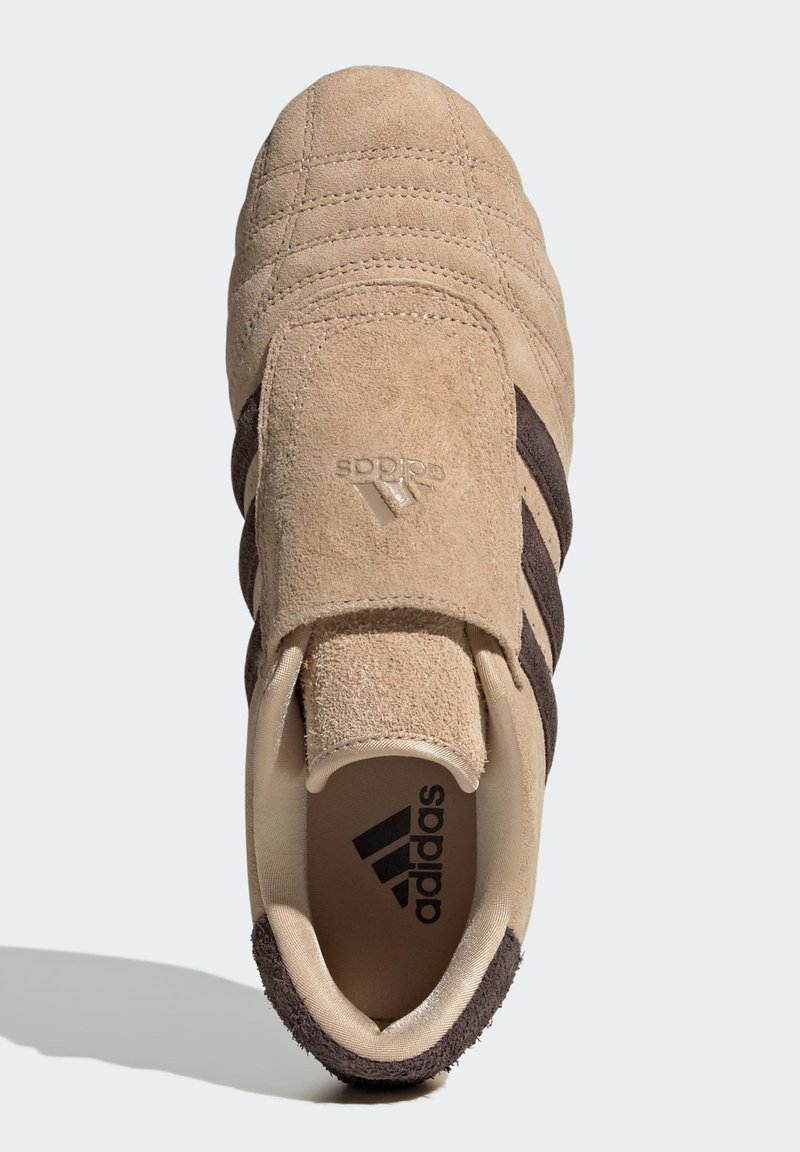 Zapatillas de ante beige con acentos y franjas en marrón oscuro. Presentan una lengüeta acolchada y una superficie texturizada. Marca en el lateral y la lengüeta.
