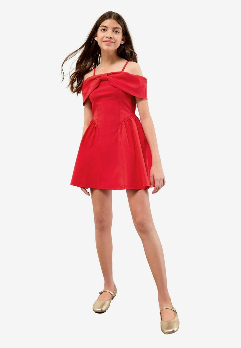 Rød off-shoulder kjole med en sløjfedetalje ved halsen. Udflydende skørt, glat tekstur. Sammen med gyldne balletflade.