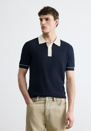 Jeune homme portant un polo bleu marine à manches courtes avec un col blanc et un jean beige, debout, une main dans la poche, regardant vers la gauche.