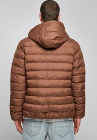 Bruine pufferjas met horizontale quilt, een capuchon en een gladde, glanzende stof. Het ontwerp is vierkant met een aansluitende taille, gecombineerd met lichtblauwe spijkerbroek.