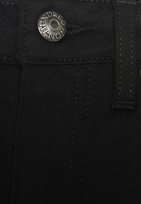 Tejido de mezclilla negro con un botón plateado visible que presenta texto grabado, y costuras decorativas a lo largo de la cintura y los bolsillos.