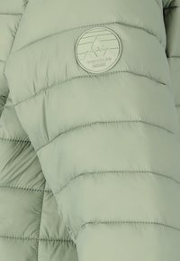 Ljusgrön pufferjacka med horisontell quiltad textur, en cirkulär logotyp på axeln och slätt, glänsande tyg.