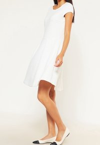 Weißes knielanges Kleid mit strukturierter Oberfläche, kurzen Ärmeln und enganliegendem Oberteil, das in einen plissierten Rock ausläuft. Kombiniert mit zweifarbigen Ballerinas.