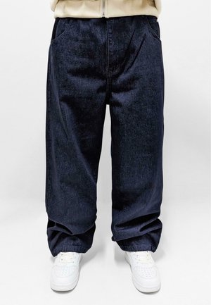 Person iført løstsiddende mørkeblå cowboybukser og hvide sneakers, der står mod en ensfarvet lys baggrund.