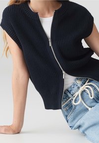 Person i svart ärmlös zip-up stickad väst över en vit skjorta och ljusblå jeans med ett vitt repbälte, lutad mot sin arm.