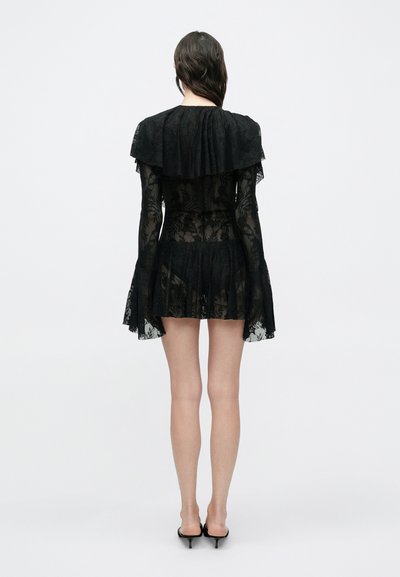 Γυναίκα που στέκεται με την πλάτη προς τον θεατή, φορώντας μαύρο μίνι φόρεμα από δαντέλα με φαρδιά μανίκια και ψηλοτάκουνα, πάνω σε λευκό απλό φόντο.