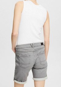 Osoba w białej bezrękawnikowej bluzce i mankietowych szarych szortach dżinsowych, pokazana od tyłu na jednolitym tle.