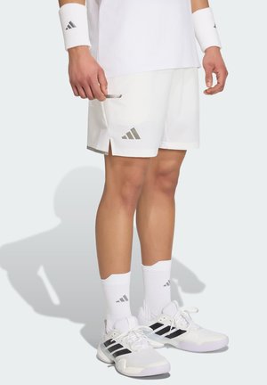 Osoba nosząca białe sportowe spodenki Adidas, opaski na nadgarstki, skarpetki oraz białe buty sportowe Adidas z czarnymi paskami, stojąca na jasnym tle.