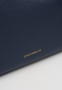 Granatowa skórzana torba o teksturowanej powierzchni i złotym logo marki "Coccinelle" na dolnej krawędzi. Prostok�ątny kształt, minimalistyczny design.