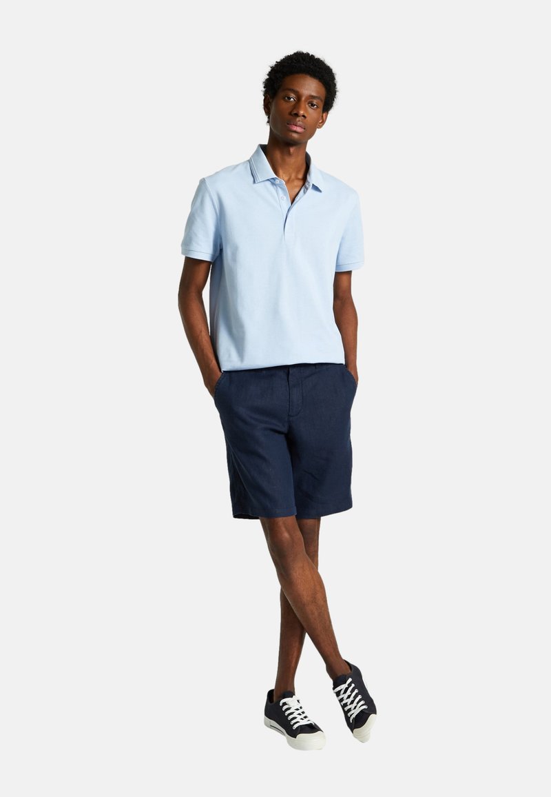 Polo bleu clair avec un col, associé à un short navy et des baskets noires avec des lacets blancs. Le modèle se tient les mains dans les poches.