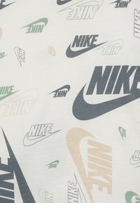 Baltas audinys su pakartotinais Nike logotipais įvairiais dydžiais ir blankiomis spalvomis: žalia, smėlio, pilka ir juoda, su glotnia tekstūra.