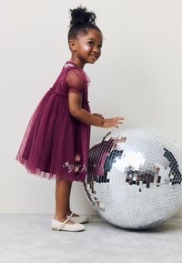 Burgunderröd tyllklänning med genomskinliga puffärmar, blomsterbroderier och insvängd midja; kombinerad med ljusa platta skor bredvid en spegelklädd discokula.