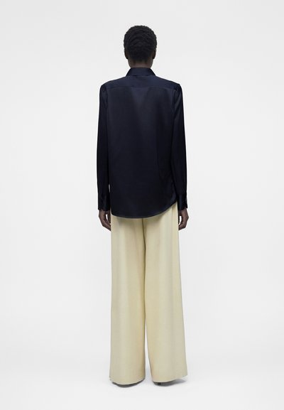 Άτομο που στέκεται με την πλάτη γυρισμένη, φοράει μαύρο μακρυμάνικο πουκάμισο και μπεζ φαρδιά παντελόνια σε απλό ανοιχτό φόντο.