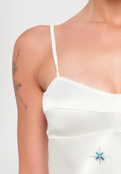 Witte satijnen camisole met dunne bandjes, voorzien van een delicate kanten rand en een blauwe sterrenvormige versiering aan de onderkant links.