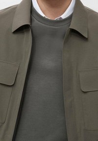 Veste vert olive avec deux poches avant, superposée sur un pull gris à col rond. Texture lisse, design simple, et ouverture frontale sans boutons.