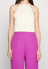 Ärmlös beige cropped topp med hög halsringning och tunna axelband. Kombinerad med texturerade, löst sittande lila byxor.